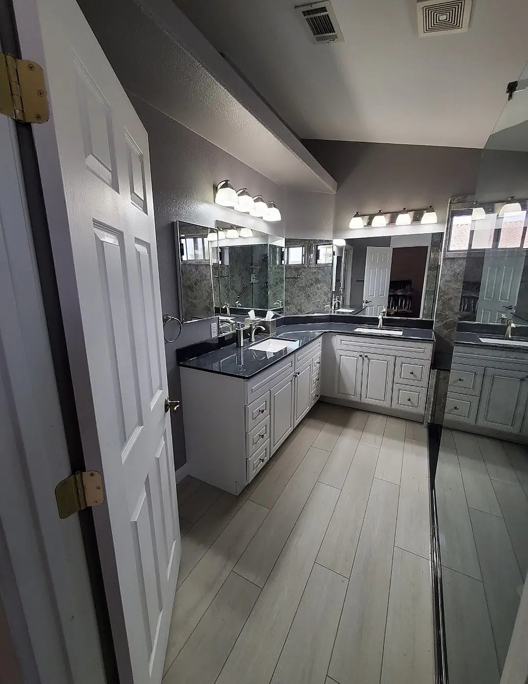 Double Vanity Master Bath — L-shaped double vanity with dark stone countertops, white cabinetry, framed mirrors and wood-look plank tile flooring, installed by Revive Bath Solutions in Athens, TX