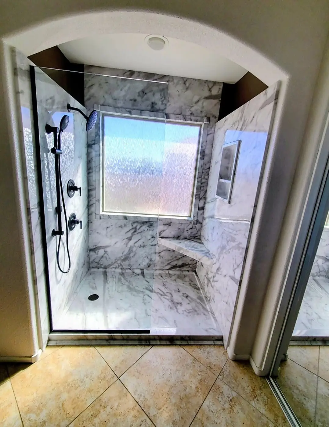 Arched Marble Walk-In Shower — grout-free marble-look wall panels with a built-in corner bench, matte black rain head and hand shower behind a frameless glass panel, installed by Revive Bath Solutions in Tyler, TX