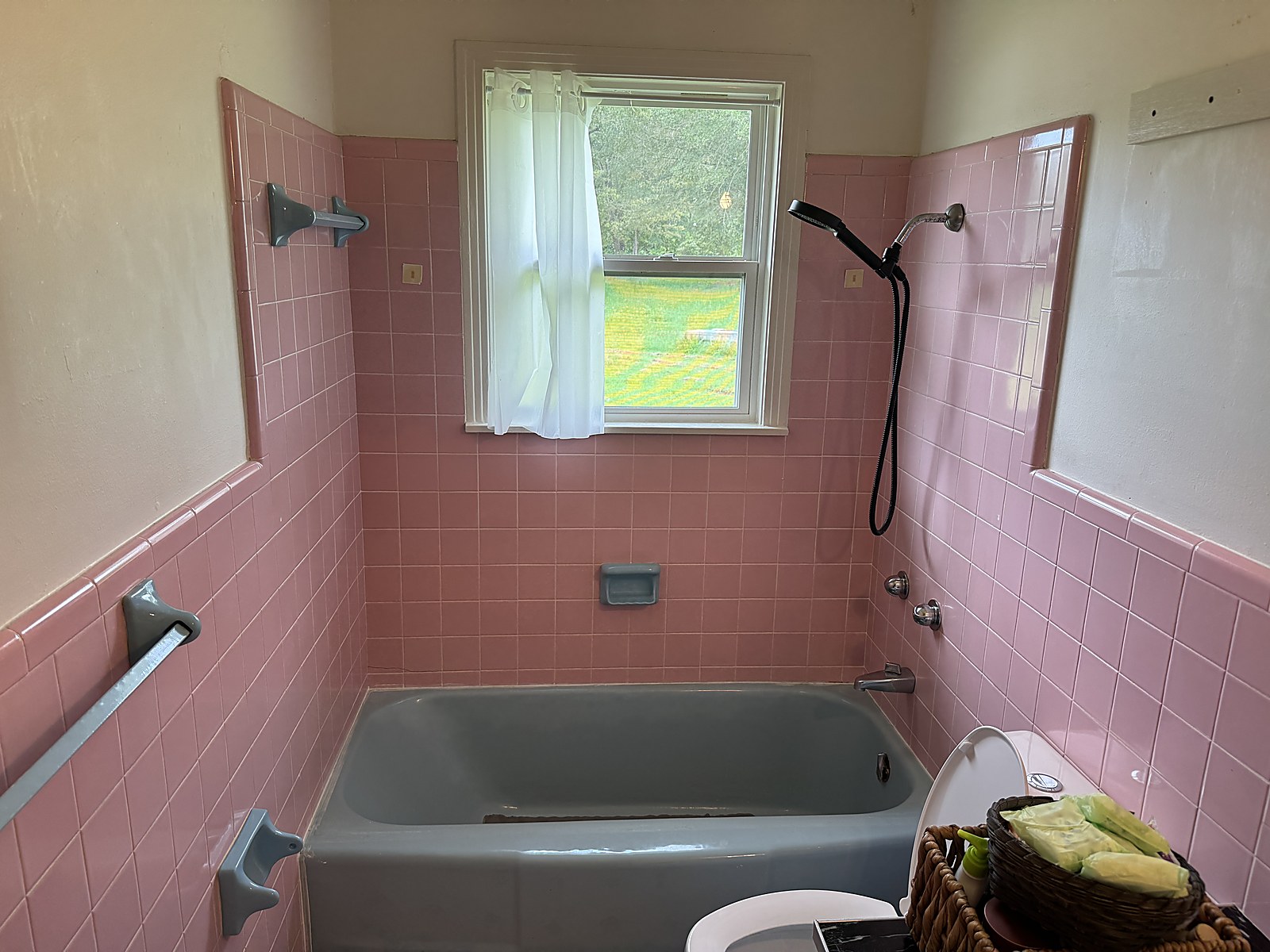 Before: original 1960s pink ceramic tile tub surround with a steel tub, grey grab bars and handheld shower in Lindale, TX